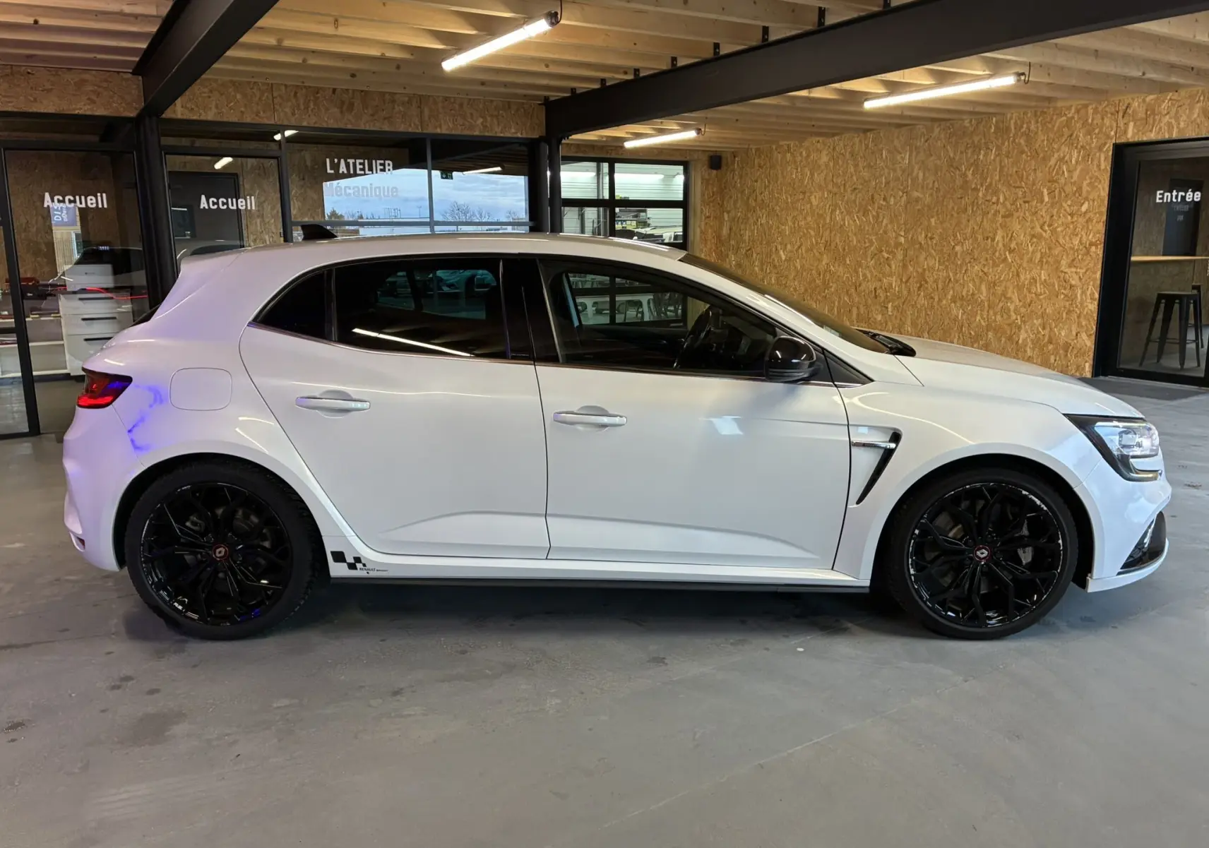 Profil côté gauche d'une Renault Mégane IV RS 280 EDC6 blanche avec jantes noires et détails sportifs, stationnée en intérieur.