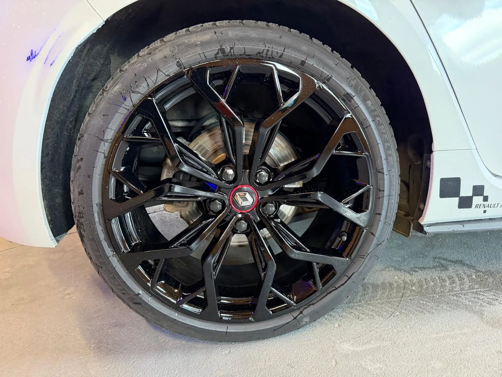 Gros plan sur la roue avant droite noire brillante d'une Renault Mégane IV RS 280 blanche avec logo rouge au centre.