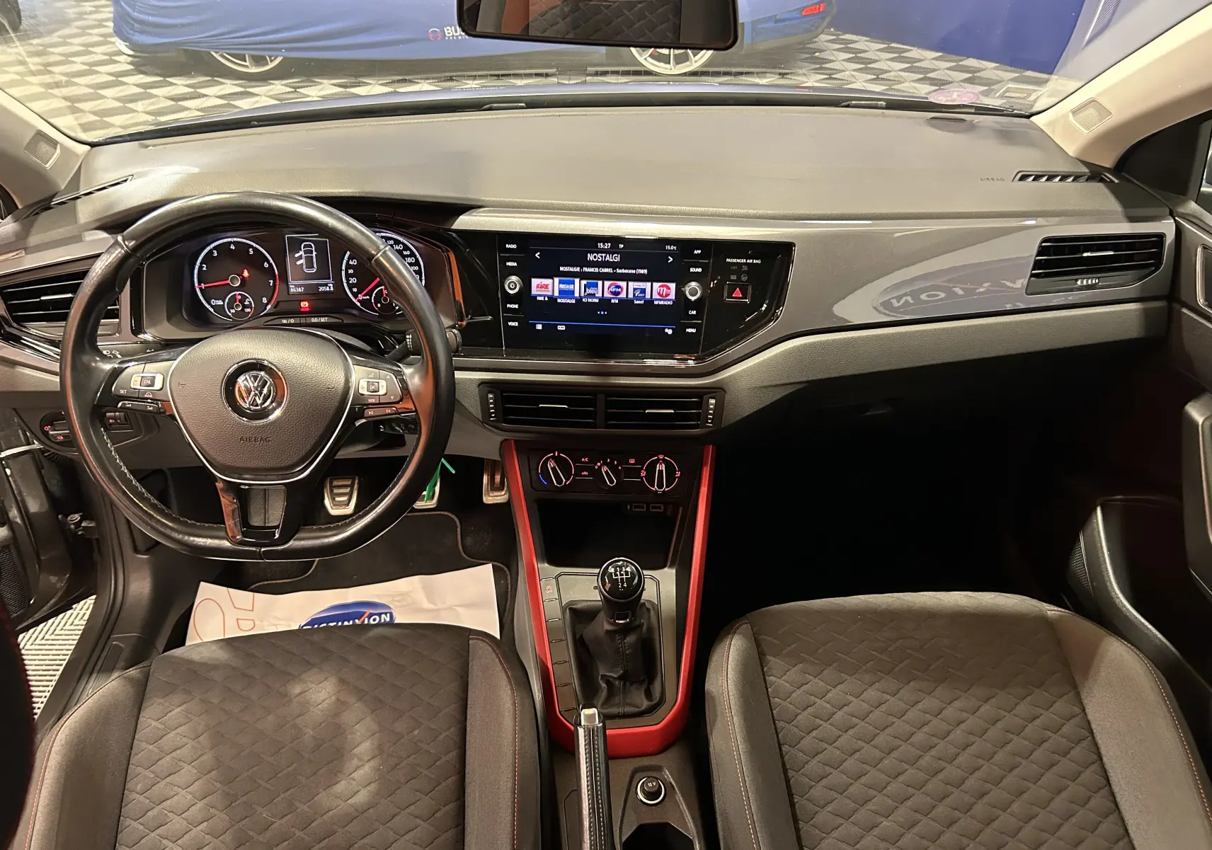 Vue intérieure avant de la Volkswagen Polo grise 2018, avec tableau de bord moderne et console centrale rouge autour du levier de vitesses manuel.