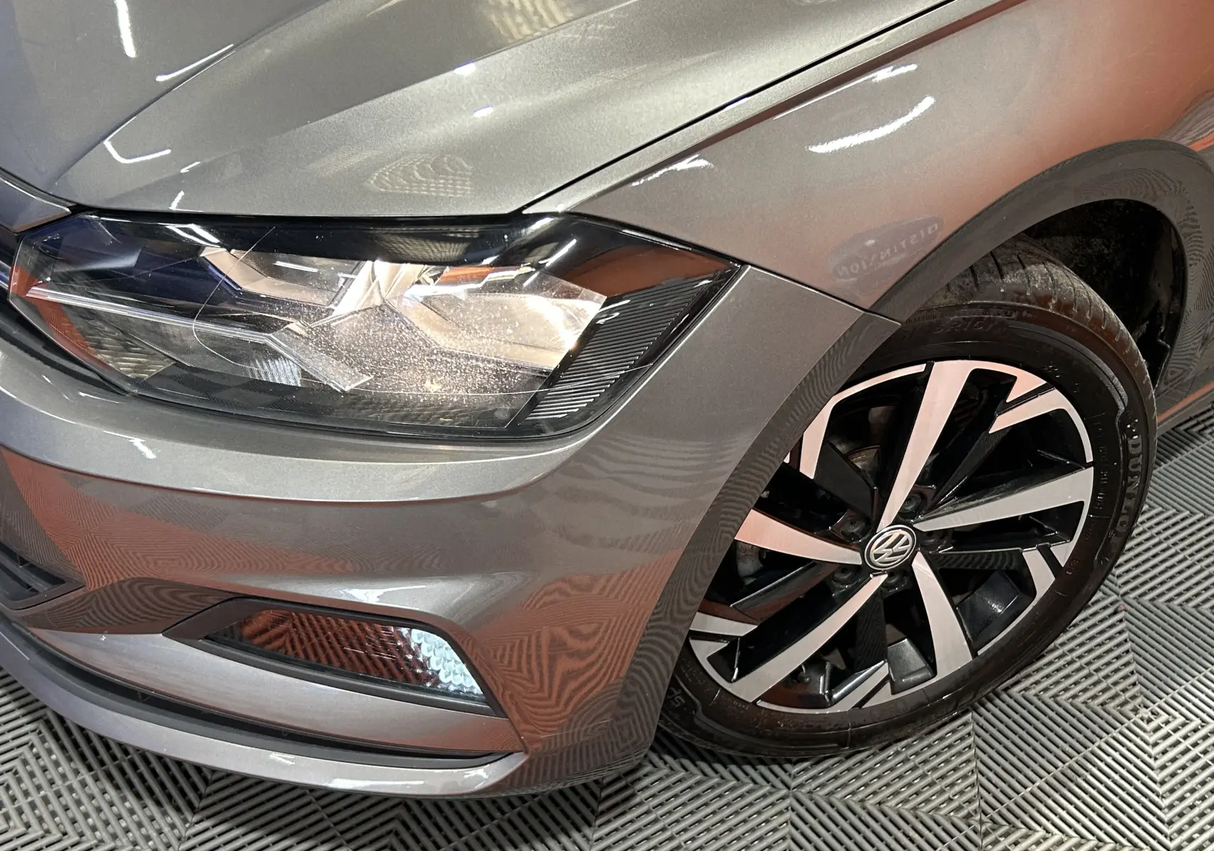 Gros plan sur l’avant droit gris métallisé d’une Volkswagen Polo 2018, mettant en valeur le phare et la jante alliage bicolore.