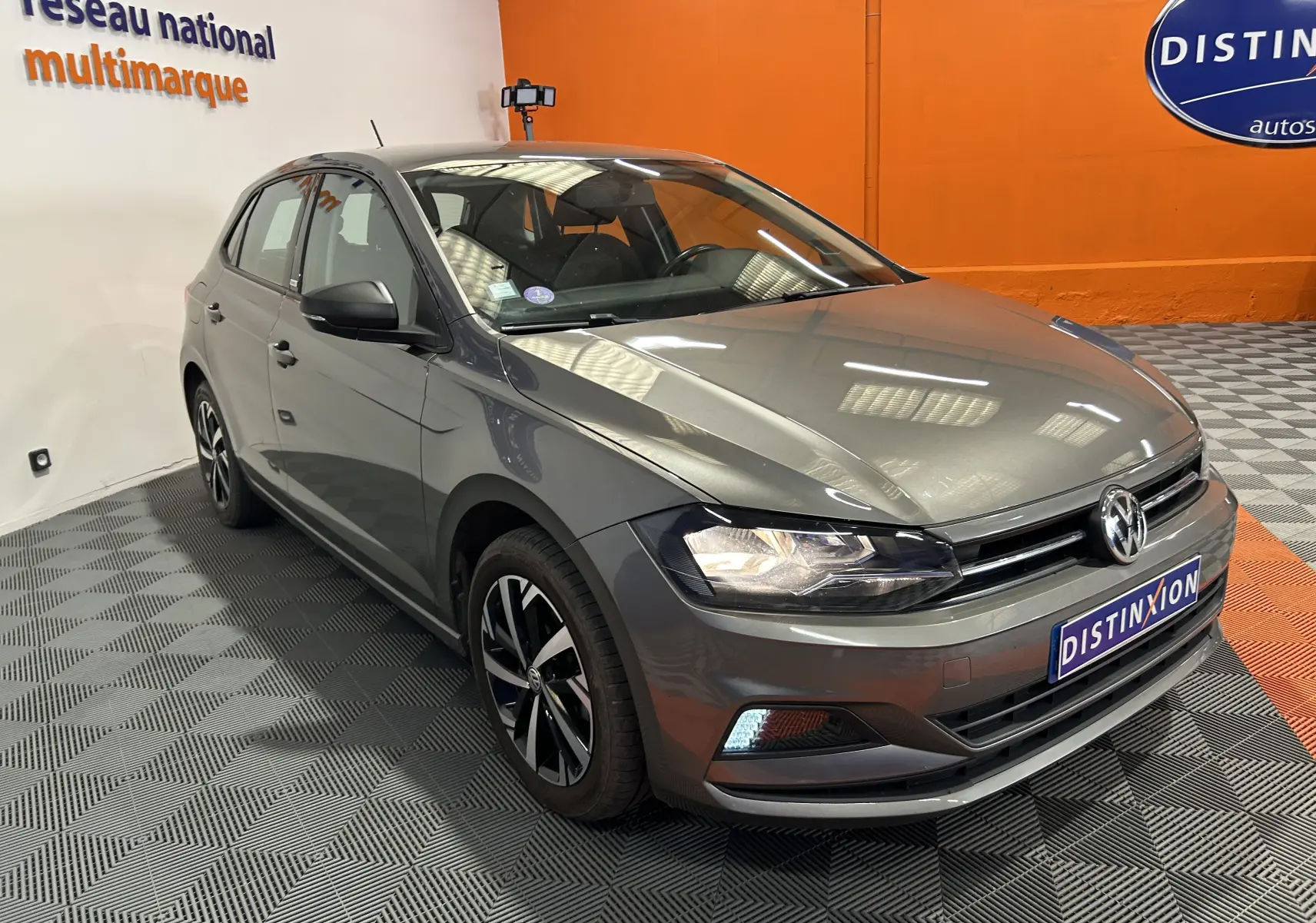 Volkswagen Polo gris vue 3/4 avant droit en intérieur avec jantes alliage et phares allumés.