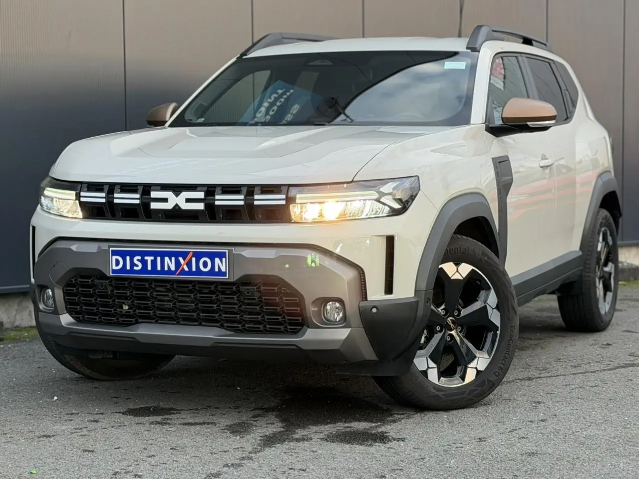 SUV Dacia Duster beige Sandstone en 3/4 avant droit, avec jantes alliage noires et phares allumés.