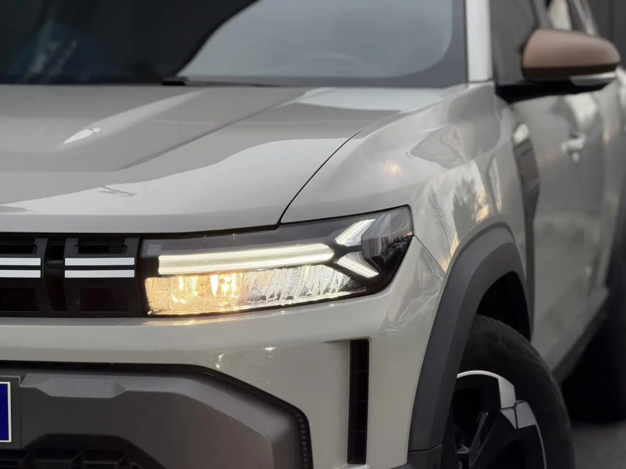 Gros plan sur l'avant droit beige Sandstone du Dacia Duster 2025, mettant en valeur son phare LED et la calandre noire.