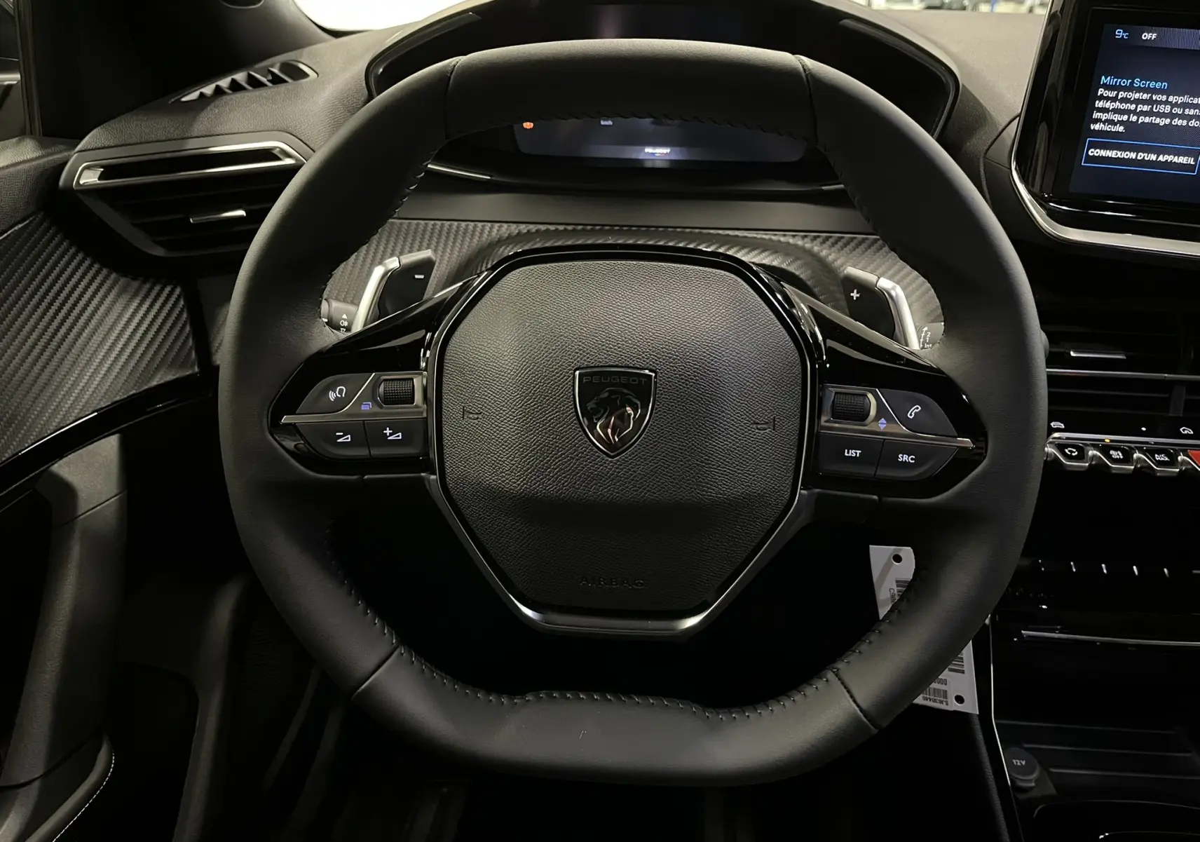 Vue rapprochée du volant noir au design hexagonal du Peugeot 2008 1.2 Hybrid 145ch, avec tableau de bord et écran tactile visibles.