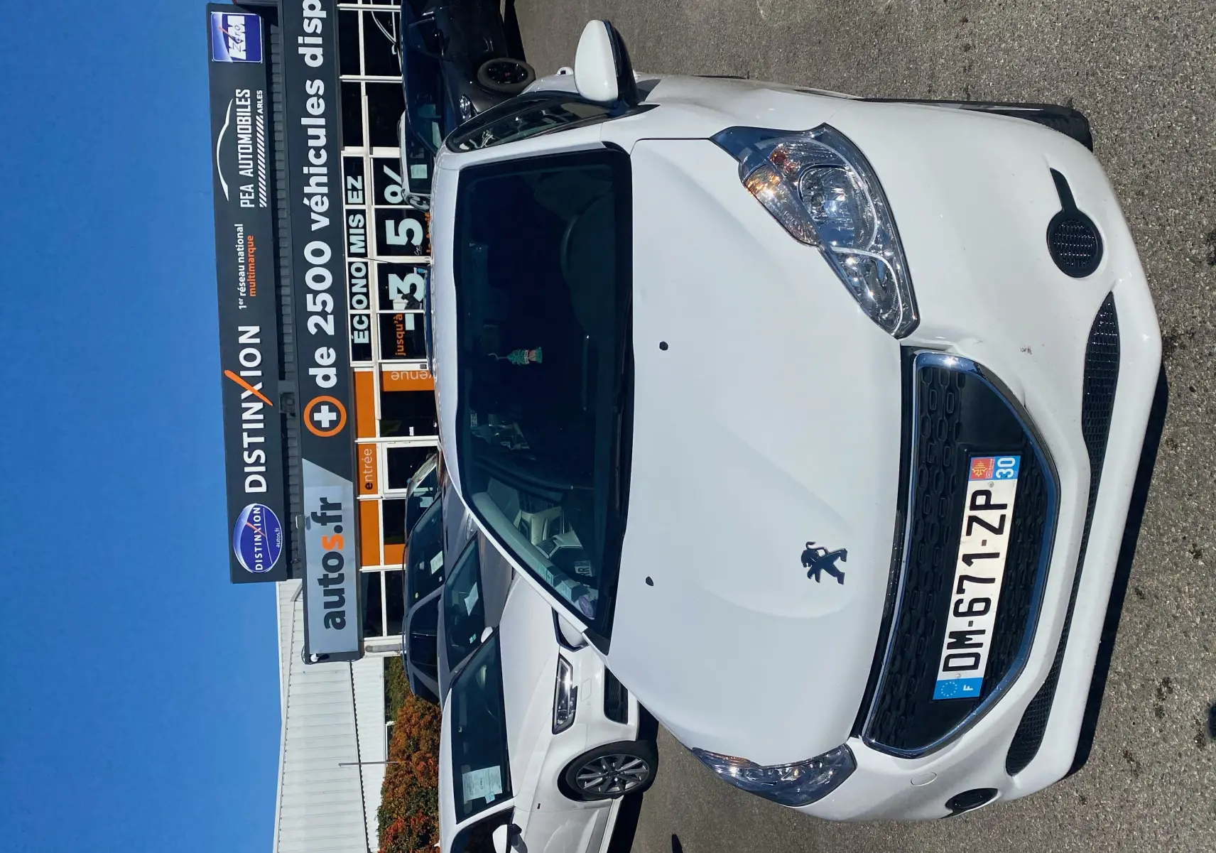 Vue 3/4 avant d'une Peugeot 208 blanche de 2014, stationnée devant un concessionnaire sous un ciel bleu.