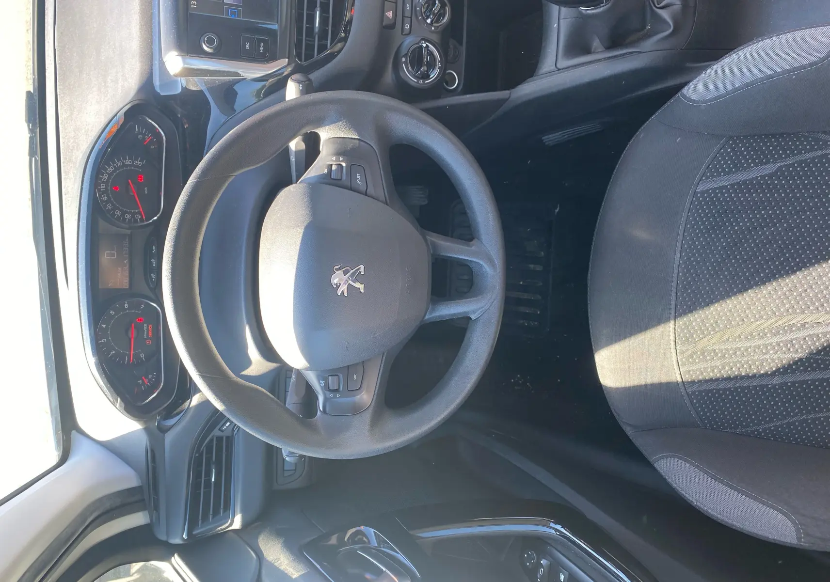 Vue intérieure centrée sur le volant et tableau de bord d'une Peugeot 208 blanche, avec sièges noirs et commandes visibles.