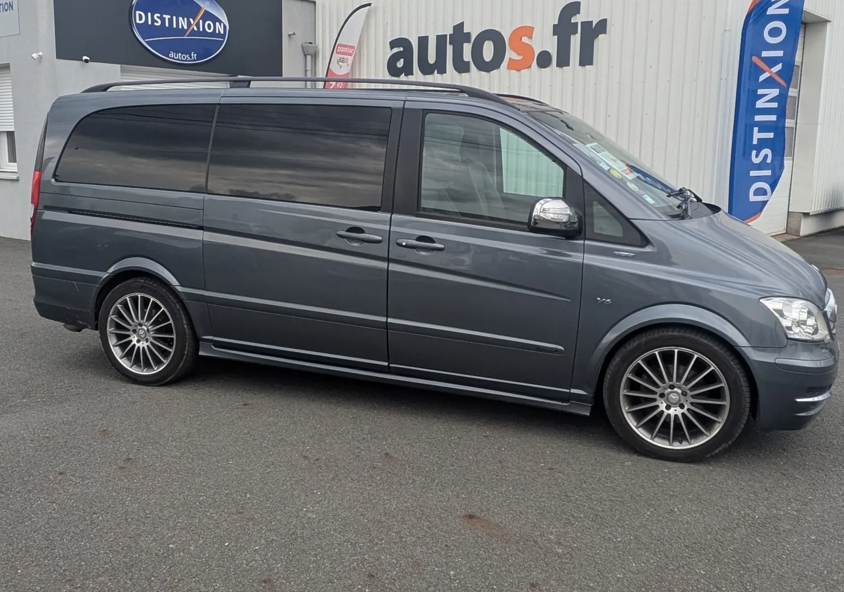 Profil côté gauche d'un Mercedes Viano gris avec vitres arrière teintées et jantes alliage devant un garage autos.fr