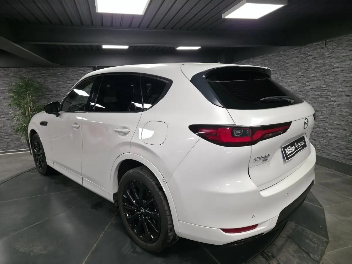 Vue 3/4 arrière droite du Mazda CX-60 blanc Arctic White avec jantes alliage noires et feux arrière LED dans un showroom.