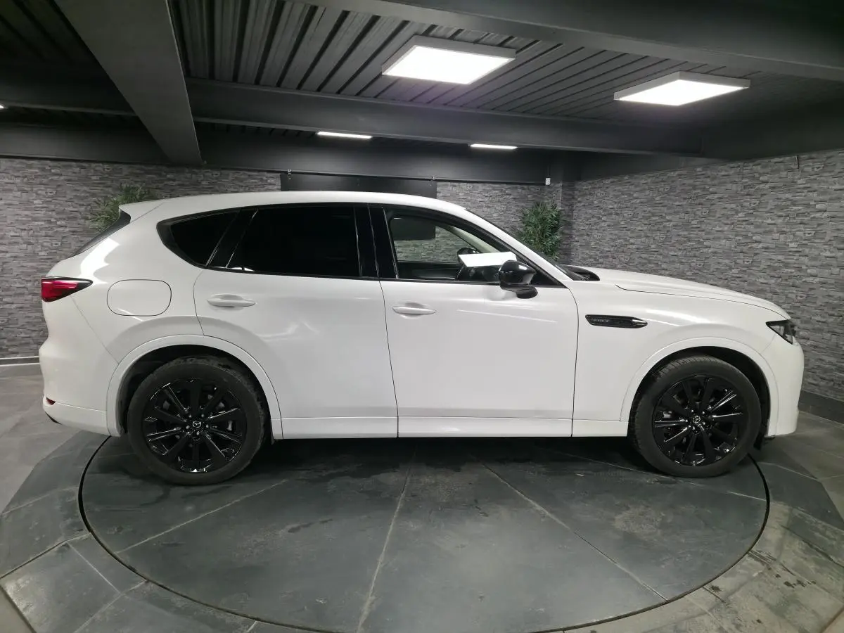Vue latérale droite du Mazda CX-60 blanc 2023 avec jantes alliage noires et vitres teintées en intérieur showroom.