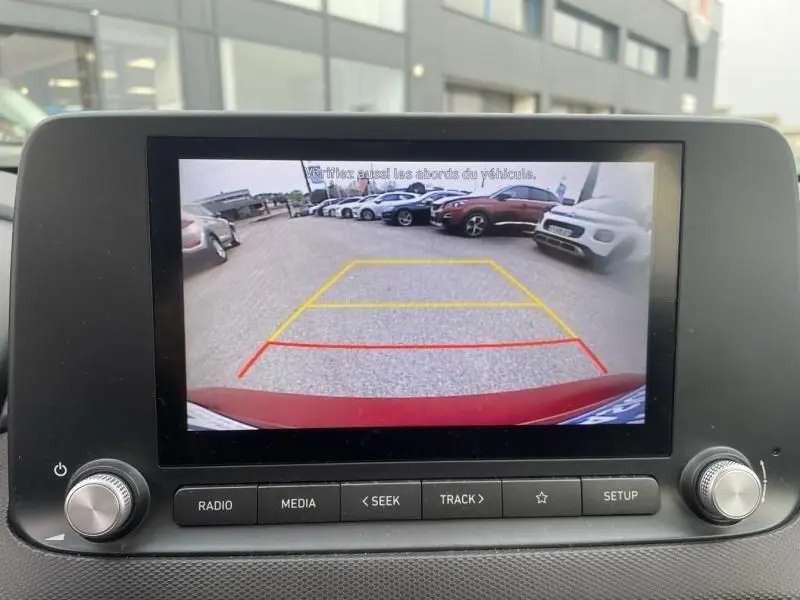 Vue arrière via caméra de recul du Hyundai Kona électrique rouge métal 2021, avec lignes d'aide au stationnement visibles.