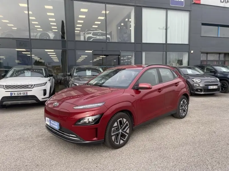 Hyundai Kona électrique rouge métal vu en 3/4 avant droit devant un showroom avec d'autres véhicules stationnés.
