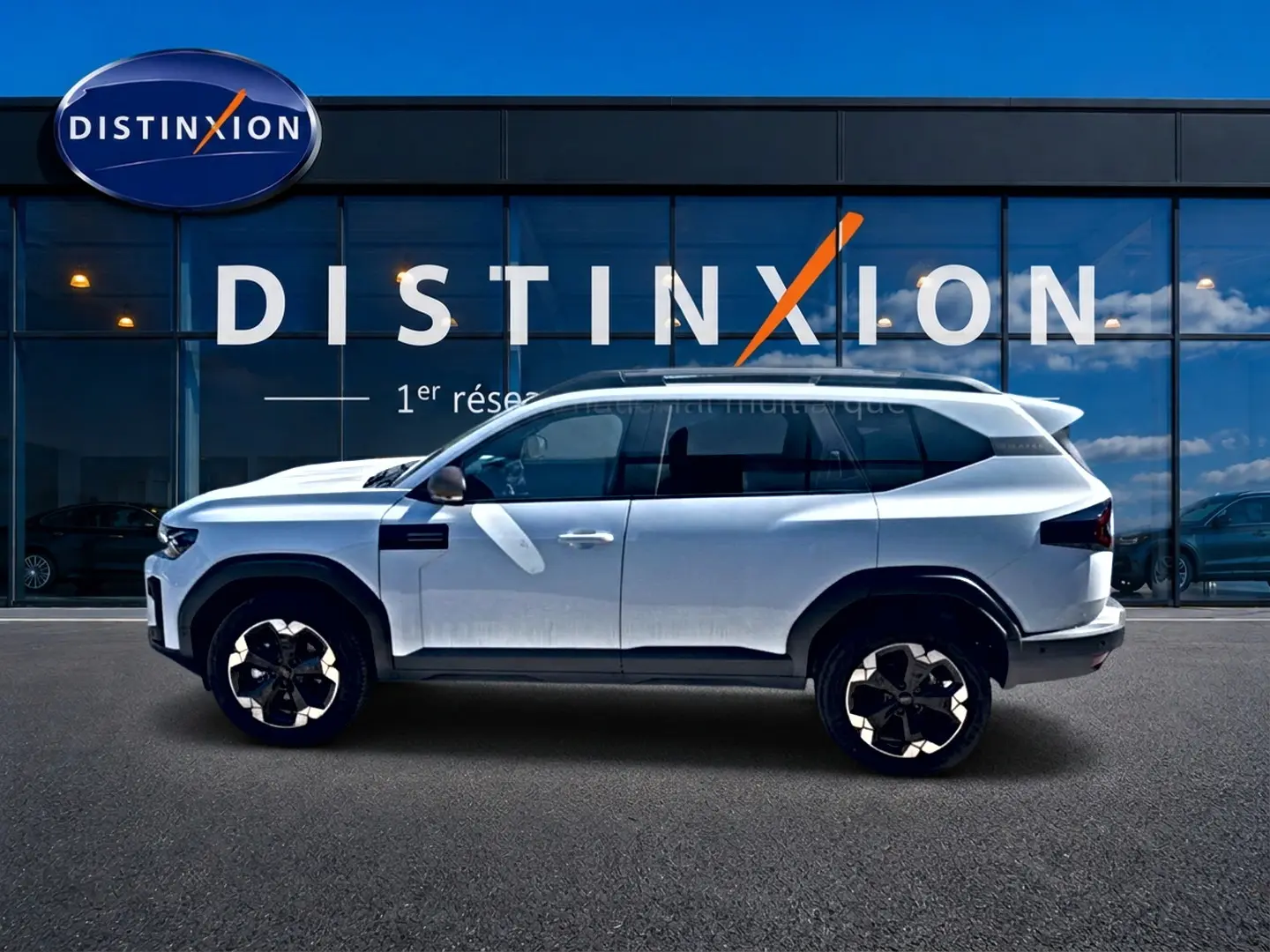 Vue de profil côté gauche du Dacia Bigster blanc 2026 avec jantes alu et barres de toit, devant une vitrine Distinxion.