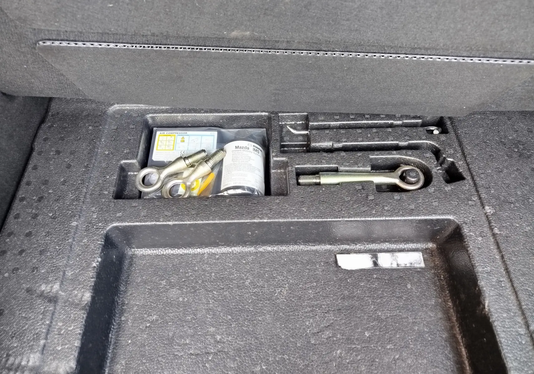 Gros plan sur le compartiment à outils du coffre du Mazda CX-30 noir, montrant les crochets de remorquage et accessoires rangés.