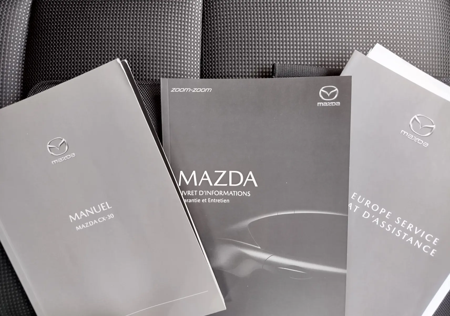 Trois manuels Mazda gris posés sur un siège en tissu noir, dont un manuel du CX-30 visible au premier plan.