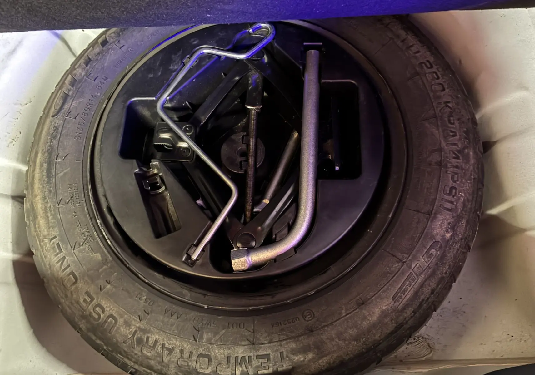 Pneu de secours noir avec outils de changement rangés au centre, vue plongeante dans le coffre d'une FIAT 500 blanche.