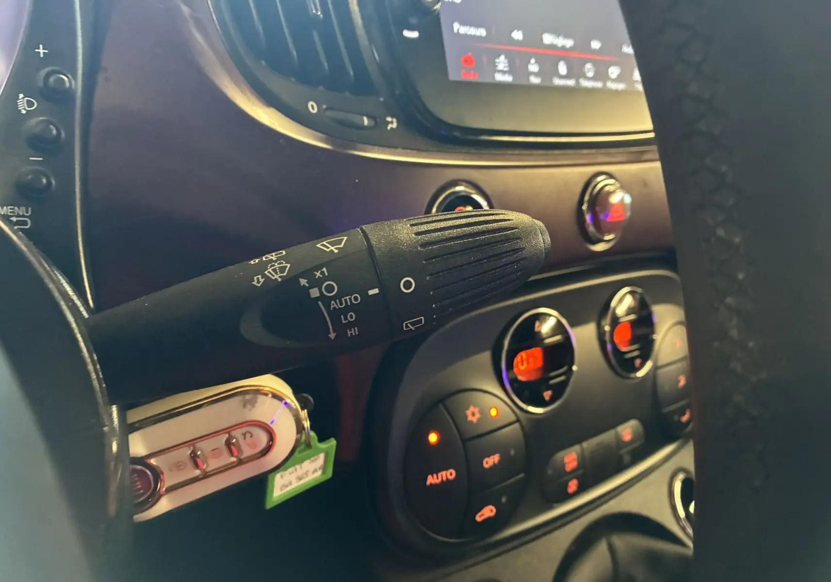 Gros plan intérieur sur la commande d’essuie-glace à gauche du volant d’une Fiat 500 blanche, avec tableau de bord et commandes clim visibles.