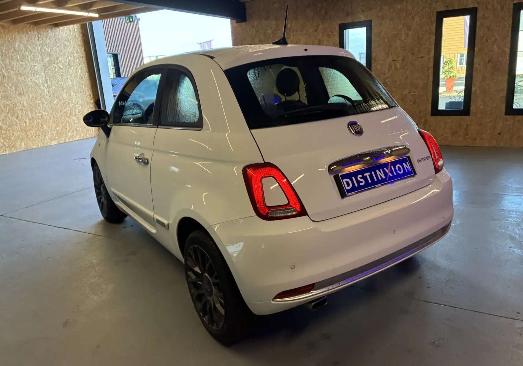 Vue 3/4 arrière droite d'une FIAT 500 blanche 2021, avec feux arrière allumés et jantes noires, stationnée en intérieur.