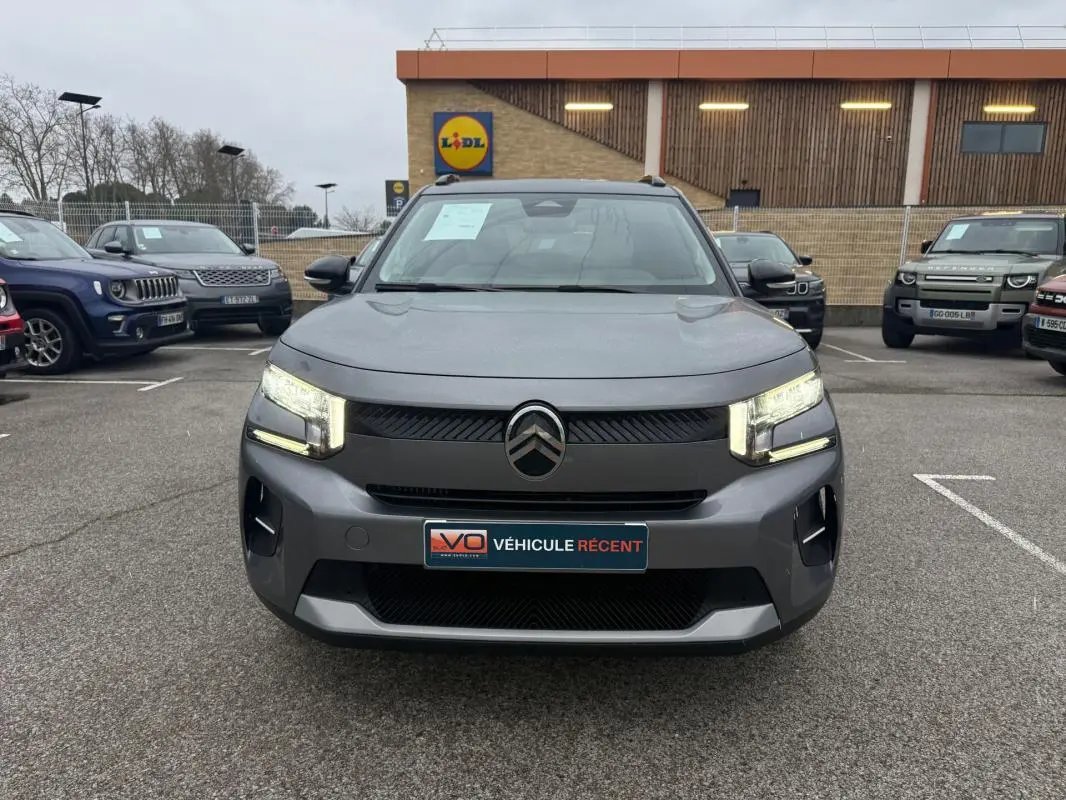 Vue avant d'une Citroën C3 gris métallisé 2025 avec phares allumés et barres de toit visibles en parking.