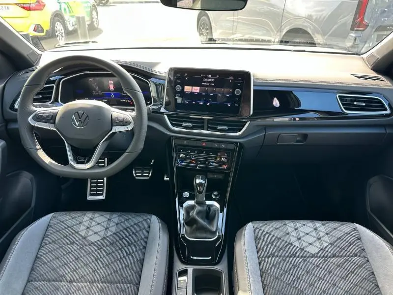 Intérieur avant du Volkswagen T-Roc 2025 avec tableau de bord noir brillant et sièges tissu gris à motifs géométriques.