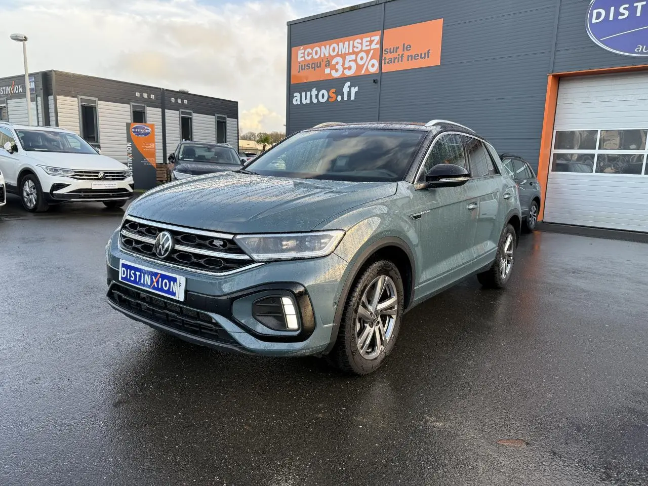 Volkswagen T-Roc bleu pétrole métallisé en 3/4 avant droit sur parking devant concession avec panneau promotionnel.