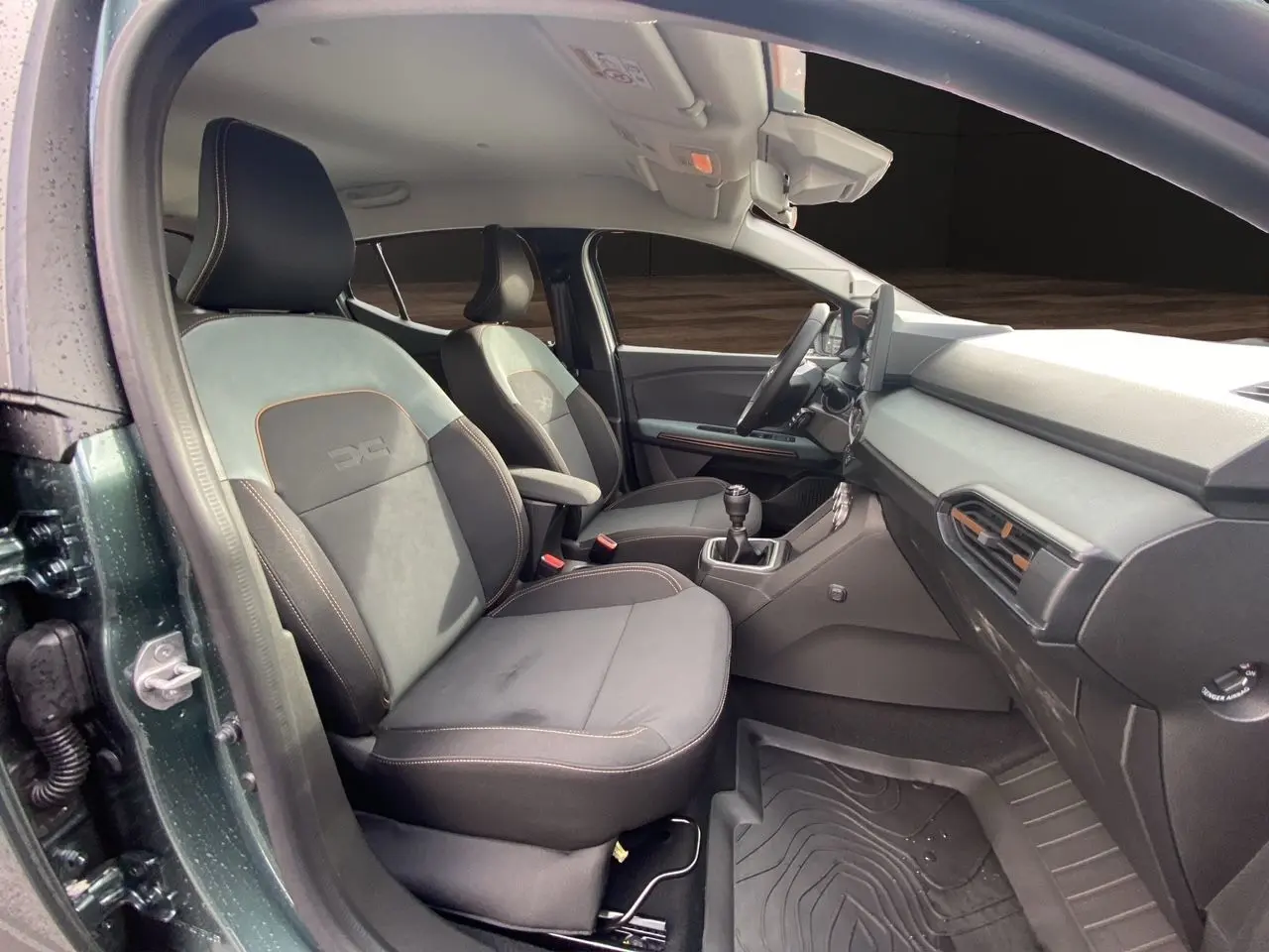 Vue intérieure côté conducteur du Dacia Sandero Stepway 2026 vert cèdre, sièges tissu gris noir avec surpiqûres orange.