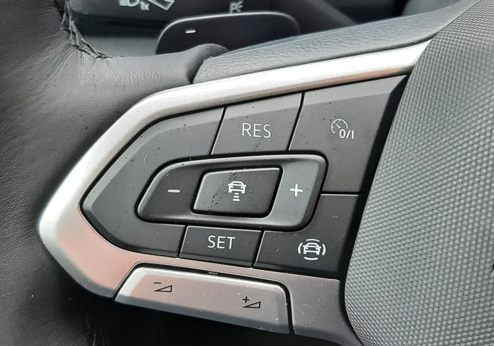 Gros plan sur les commandes au volant côté gauche du Volkswagen T-Roc gris pyrite, mettant en valeur les boutons de régulateur de vitesse.