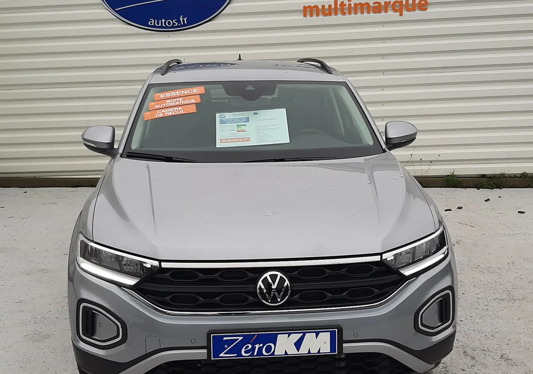 Volkswagen T-Roc gris pyrite métal vu de face, avec plaques "Zéro KM" et autocollants orange sur pare-brise.
