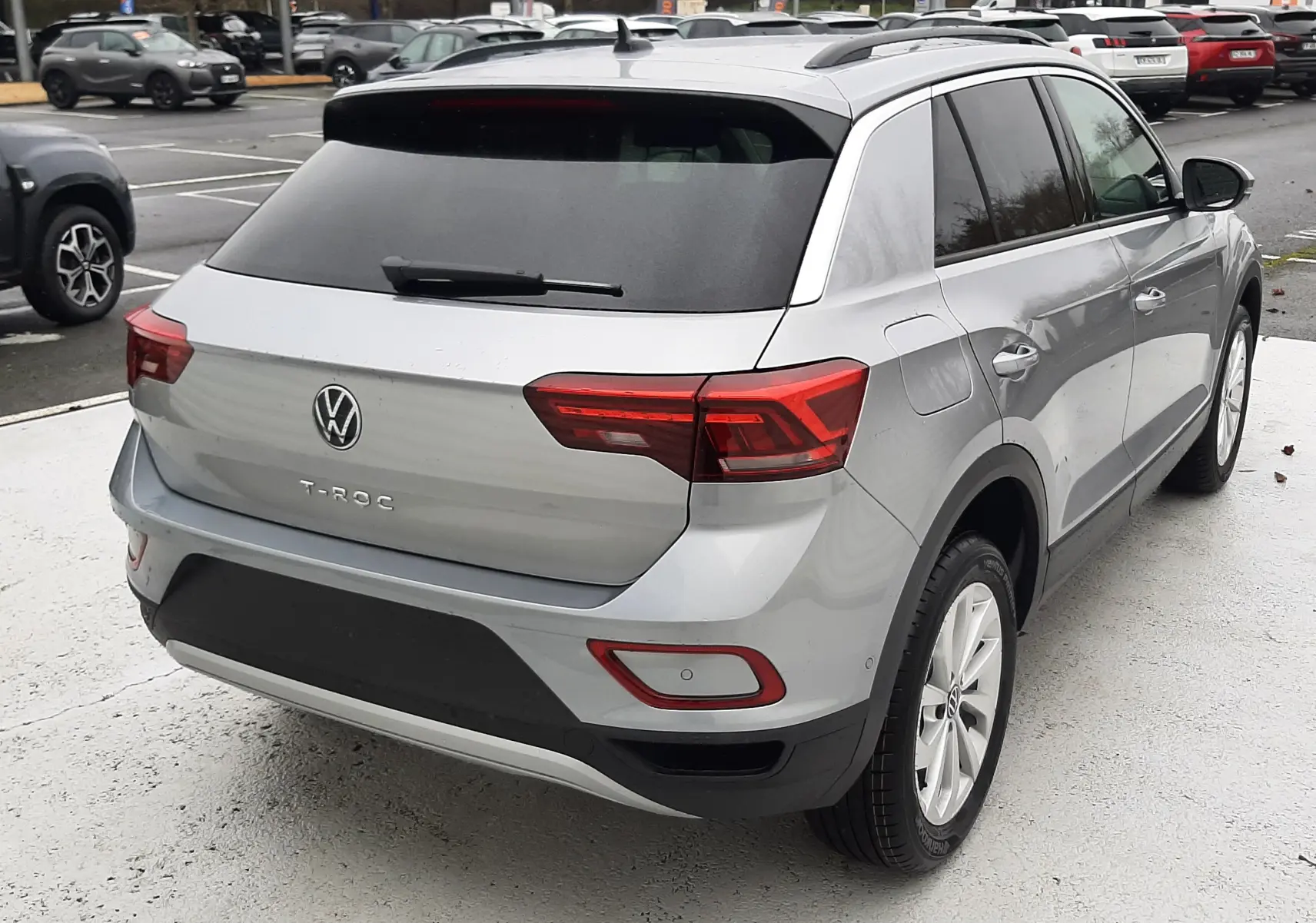 Vue 3/4 arrière droite d'un Volkswagen T-Roc gris pyrite métal 2025 avec feux arrière LED et toit noir.