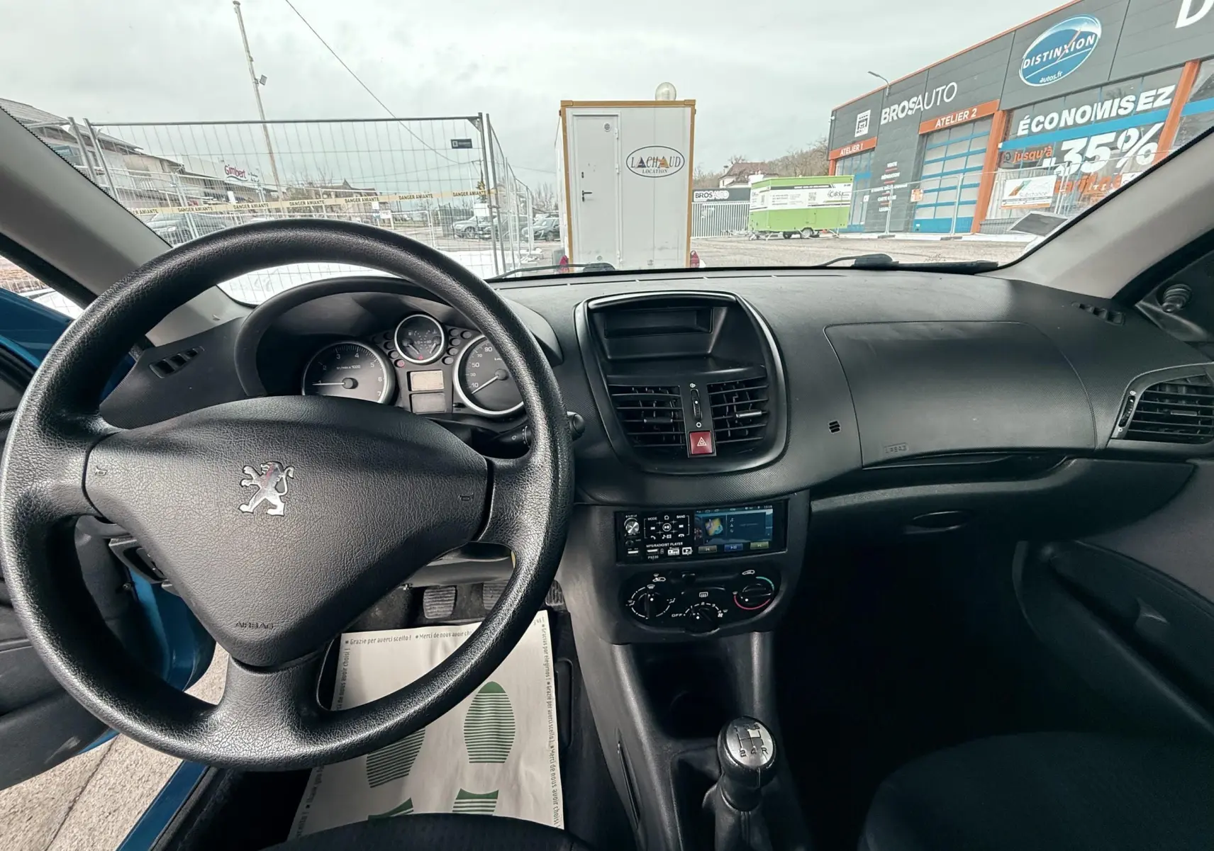 Vue intérieure avant de la Peugeot 206+ bleue, montrant le volant, tableau de bord et levier de vitesses manuel.