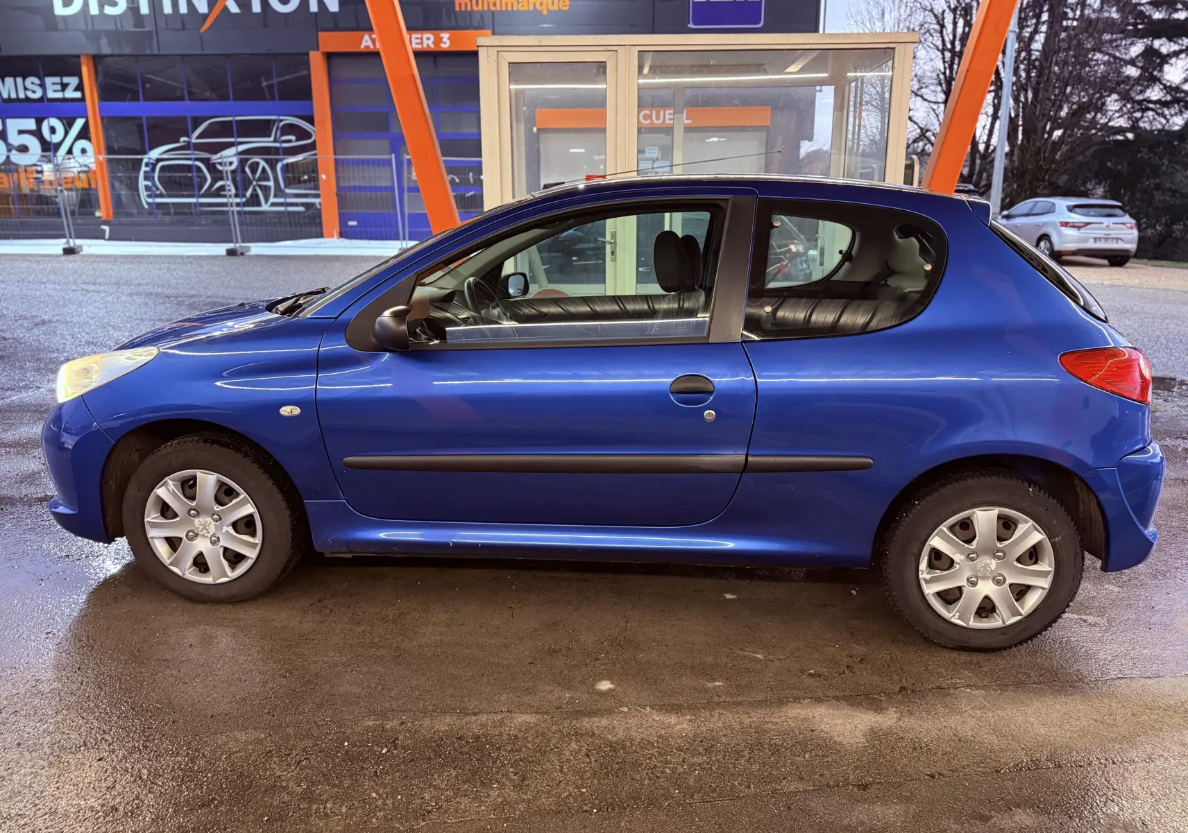 Profil côté gauche d'une Peugeot 206+ bleue à 3 portes, garée devant un bâtiment commercial.
