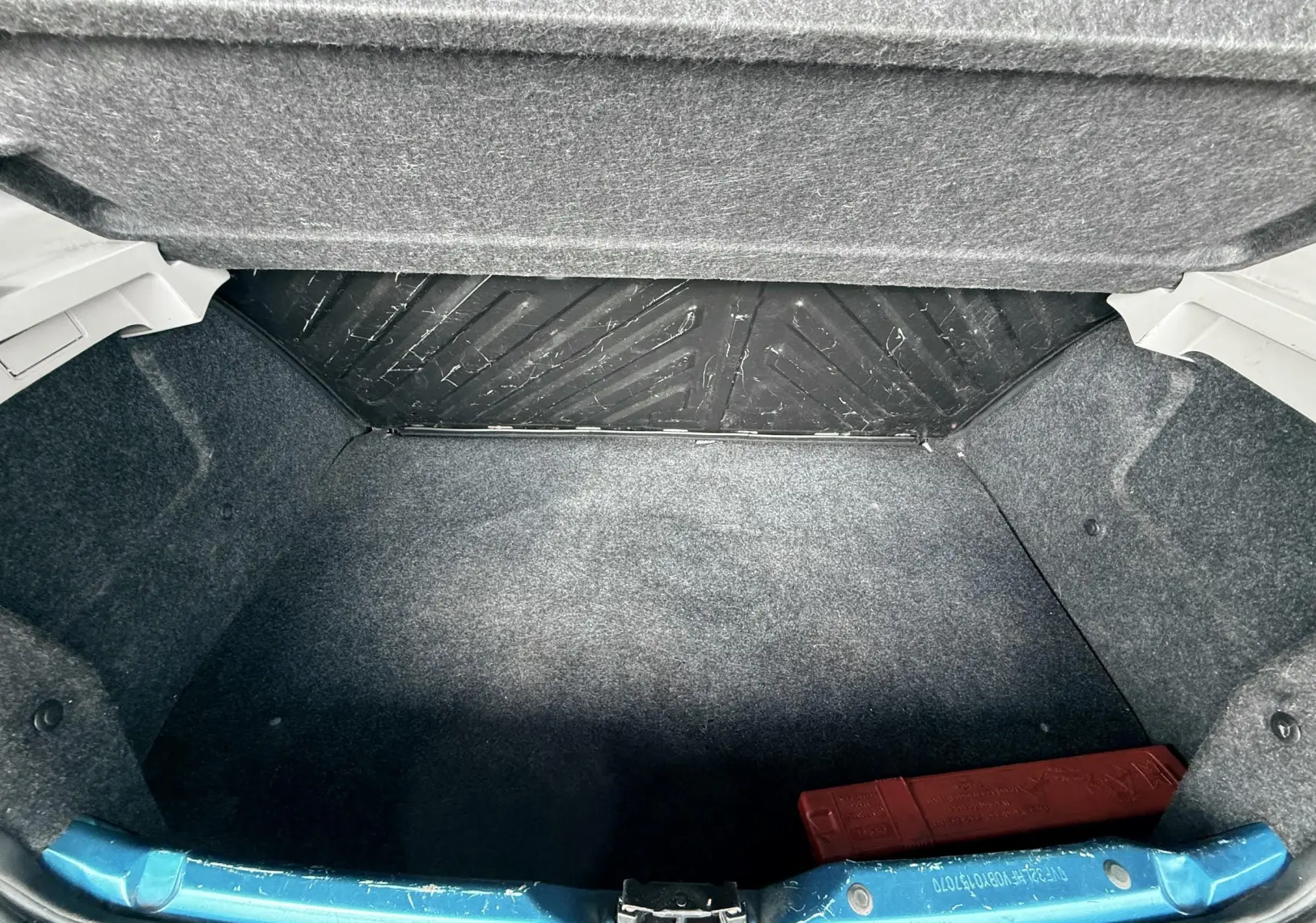 Vue intérieure du coffre ouvert d'une Peugeot 206+ bleue, montrant la garniture grise et un triangle de signalisation rouge.