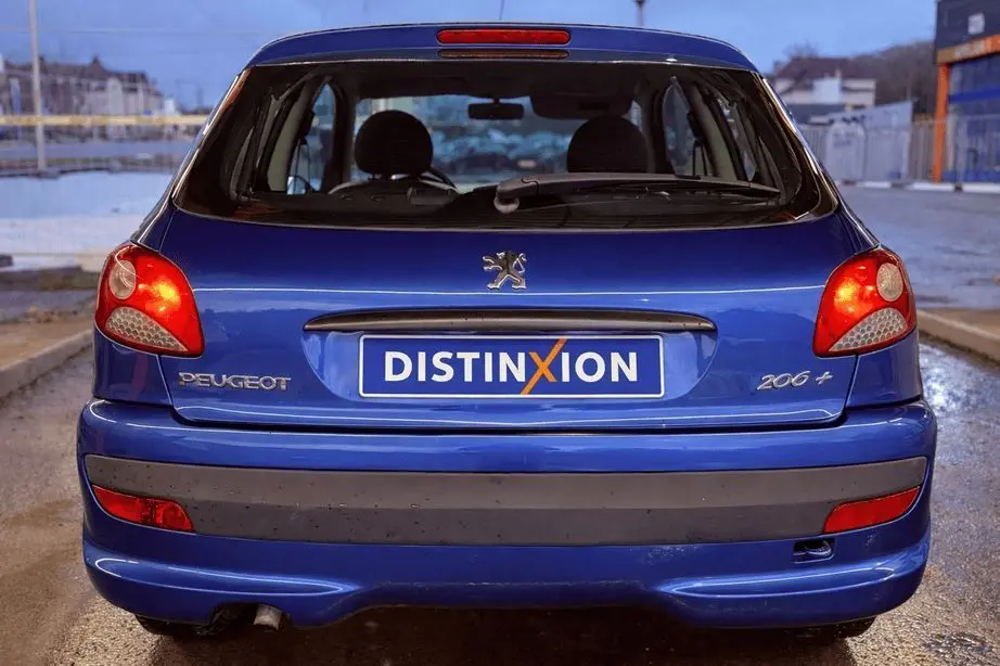 Vue arrière d'une Peugeot 206+ bleue avec plaque Distinxion, feux arrière allumés et hayon fermé.