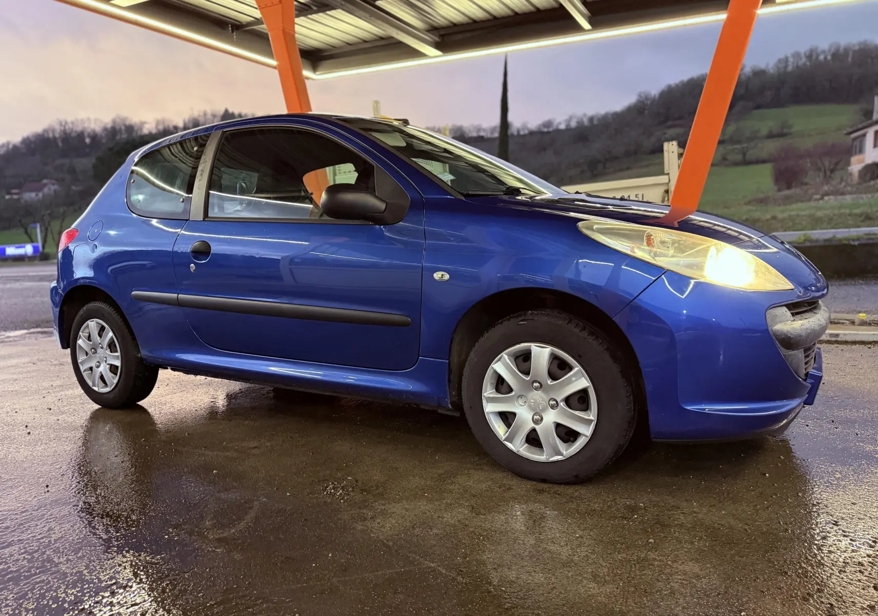 Peugeot 206+ bleu vue de profil côté gauche sous abri, phare avant droit allumé, carrosserie 3 portes.