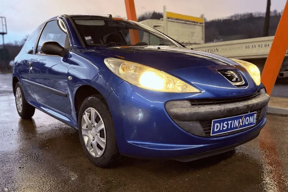 Peugeot 206+ bleu vue 3/4 avant droit avec phares allumés sur sol mouillé, calandre noire et logo visible.