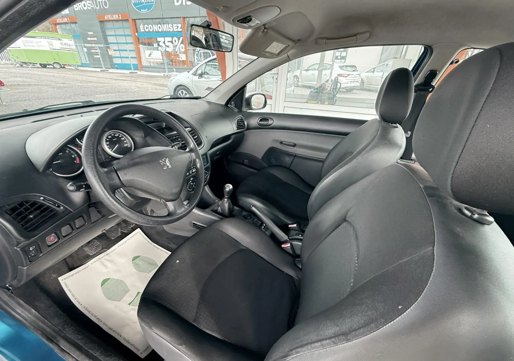 Vue intérieure côté conducteur de la Peugeot 206+ bleue, montrant le volant, la console centrale et les sièges noirs en tissu.