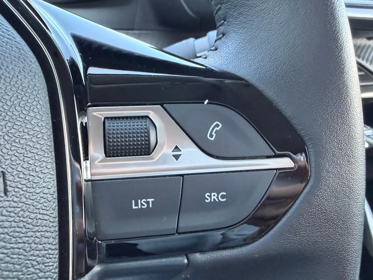 Gros plan sur les commandes audio au volant noir de la Peugeot 208 1.2 100ch Gris Artense, montrant molette et boutons SRC, LIST.