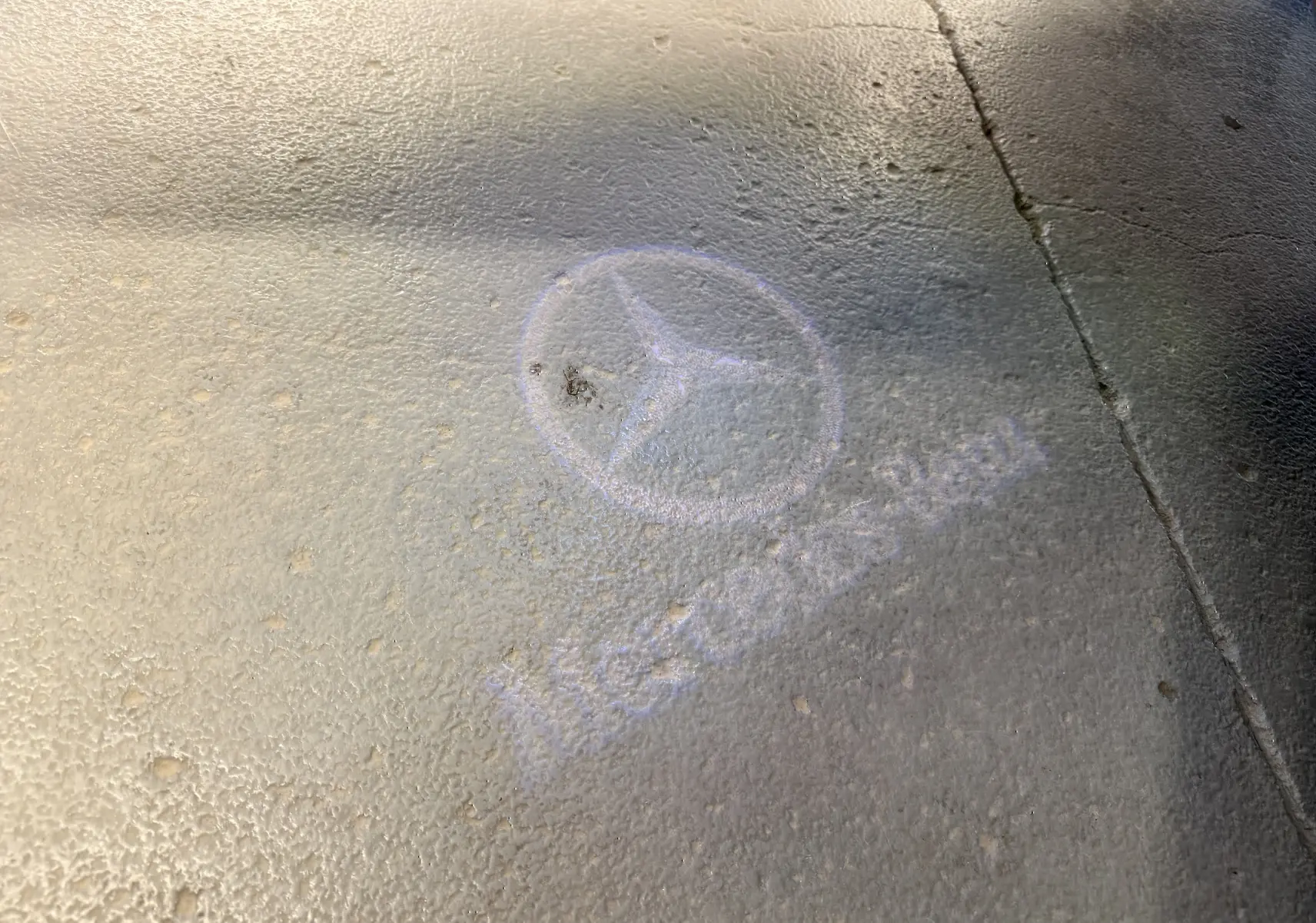 Projection lumineuse du logo Mercedes-Benz au sol sur un sol texturé, sans véhicule visible.