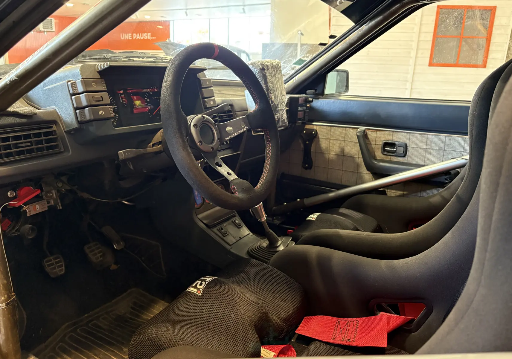 Vue intérieure côté conducteur de l'Audi Coupé GT noir de 1983, avec sièges baquets, harnais rouge et volant course.