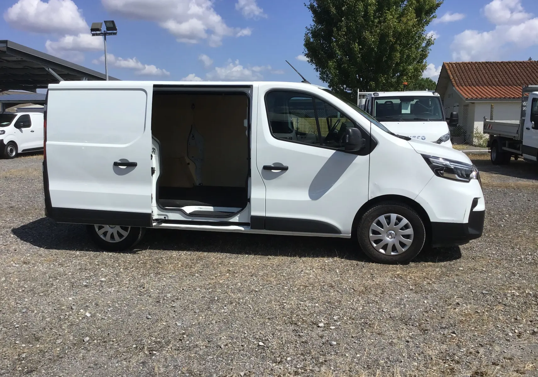 Nissan Primastar blanc vu de profil côté droit avec porte latérale coulissante ouverte sur parking extérieur.