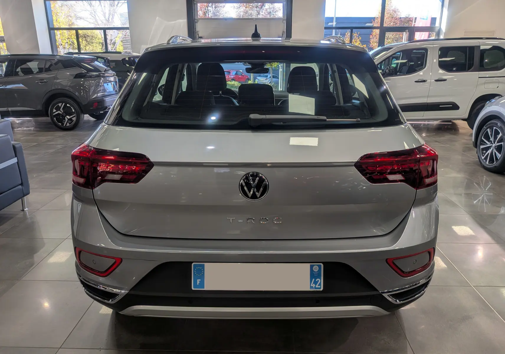 Vue arrière d'un Volkswagen T-Roc gris pyrite 2024 en showroom, avec feux arrière rouges et logo VW visible.