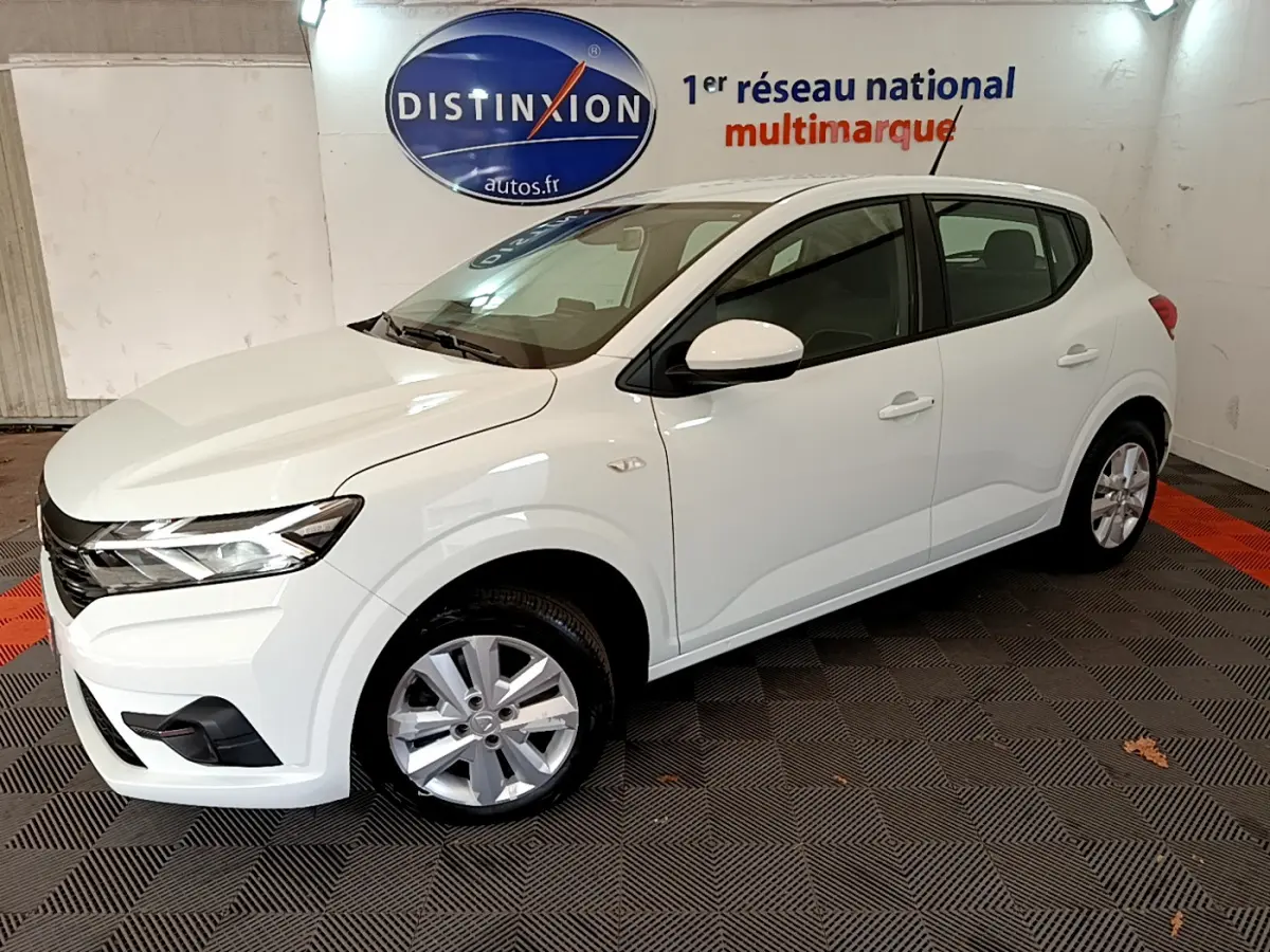 Vue 3/4 avant d'une Dacia Sandero Confort TCe 90 blanche, stationnée en intérieur sur sol damier noir et rouge.