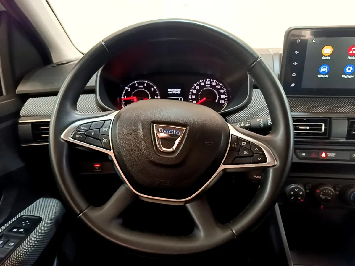 Vue intérieure centrée sur le volant cuir noir avec logo Dacia, tableau de bord et écran tactile du Sandero blanc 2022.