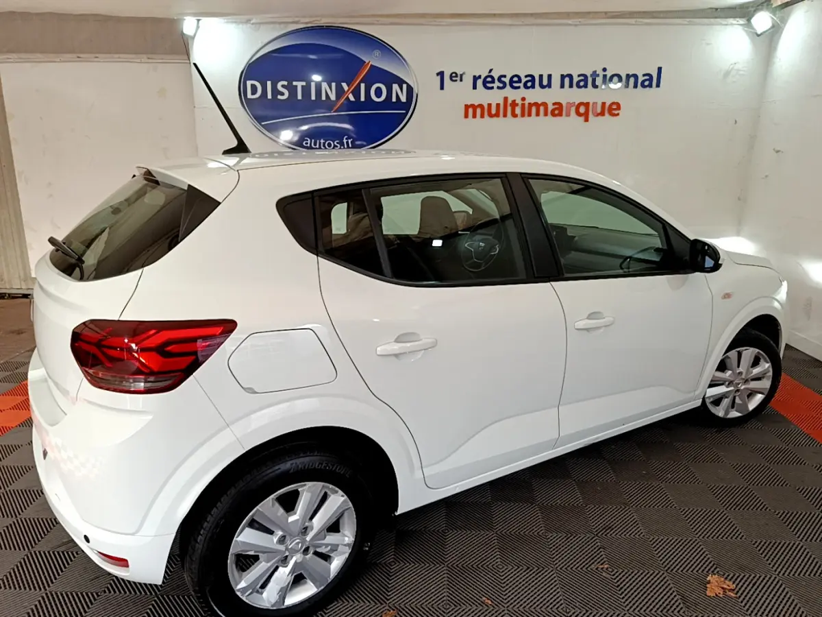 Vue 3/4 arrière droite d'une Dacia Sandero Confort blanche 2022 dans un showroom avec logo Distinxion en arrière-plan.