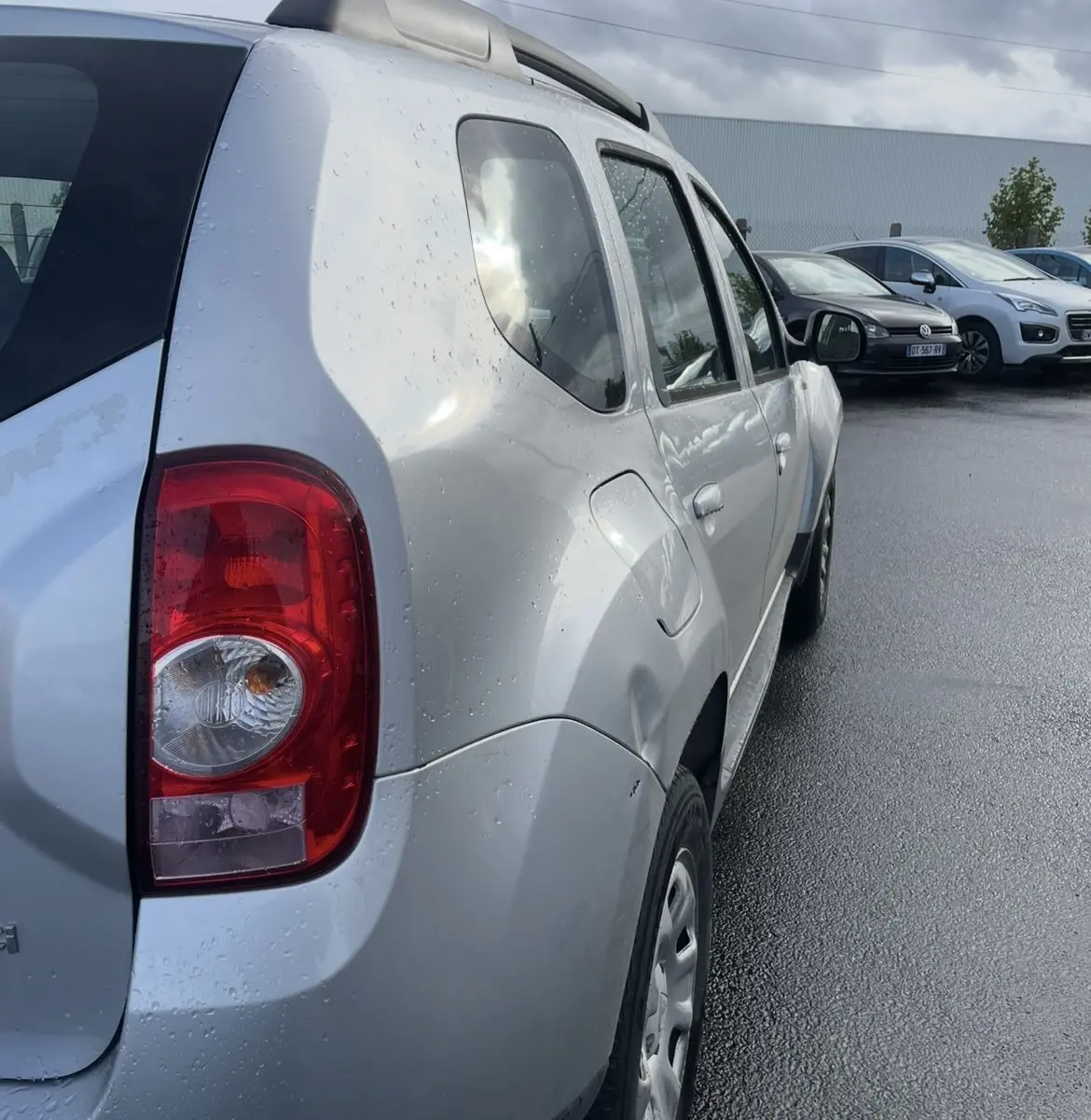 Vue 3/4 arrière gauche d'un Dacia Duster gris avec barres de toit et attelage sous un ciel nuageux.