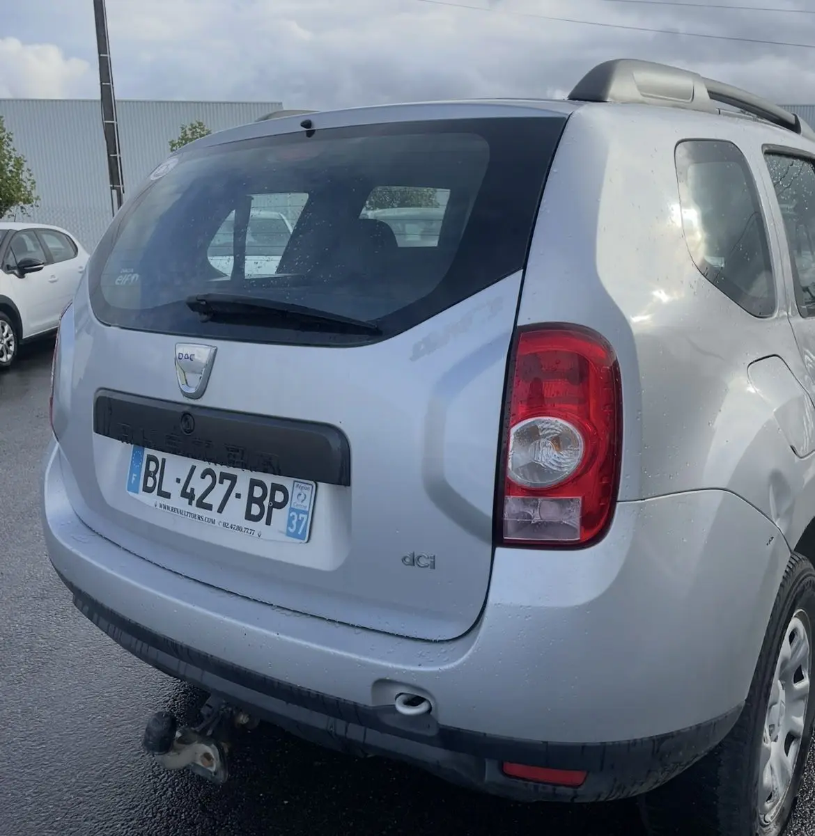 Vue arrière droite d'un Dacia Duster gris 2011 avec attelage et barres de toit sous un ciel nuageux.