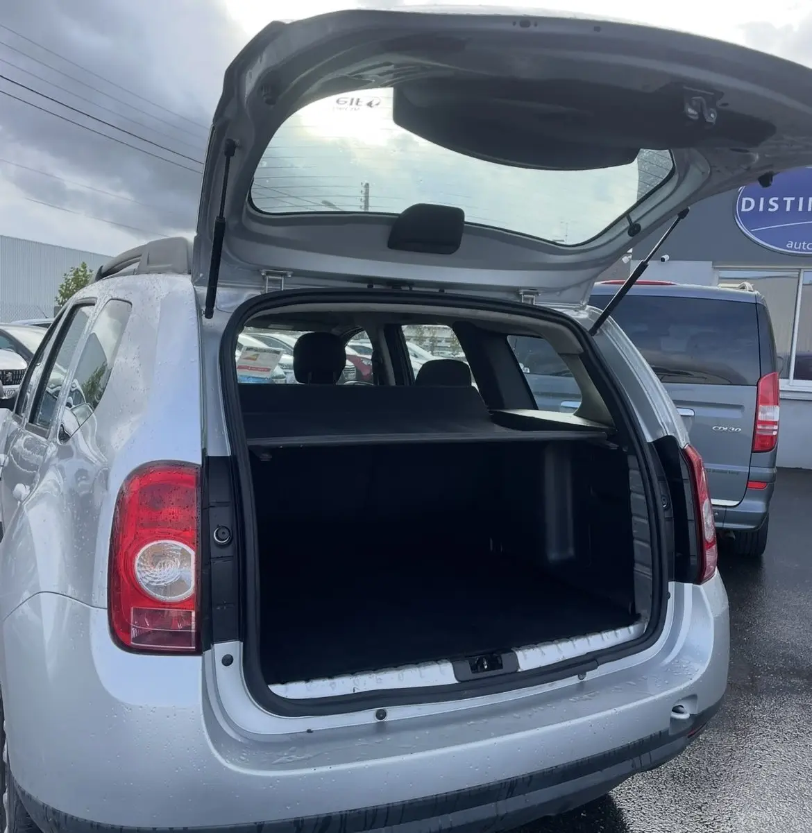 Vue arrière ouverte d'un Dacia Duster gris 2011 avec attelage et coffre spacieux visible sous un ciel nuageux.
