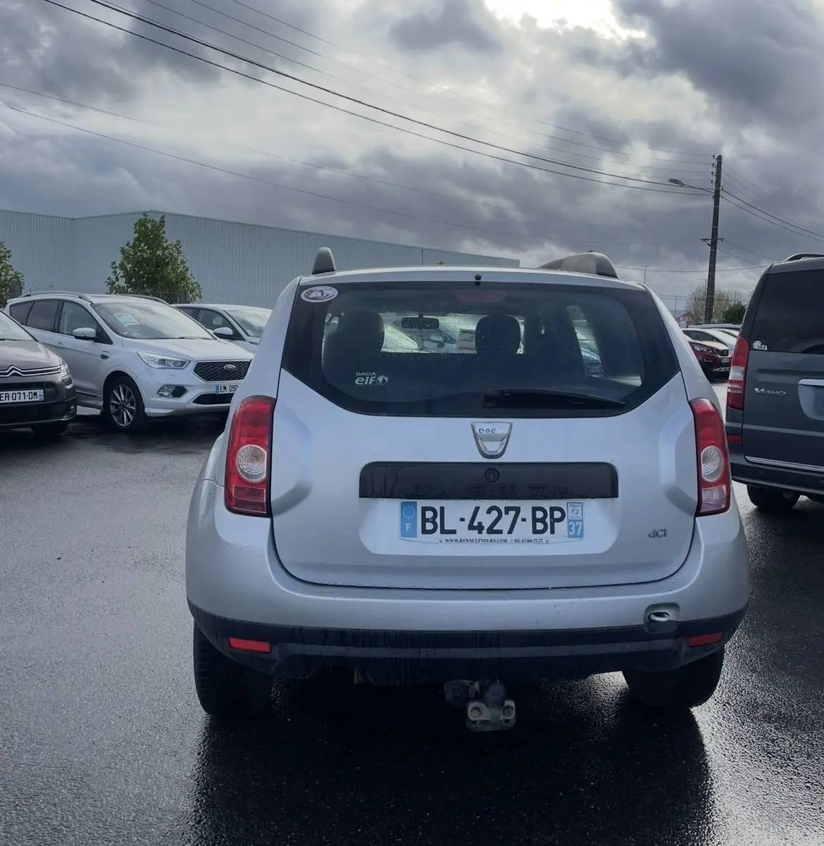 Vue arrière d'un Dacia Duster gris avec attelage et vitres teintées sous un ciel nuageux.