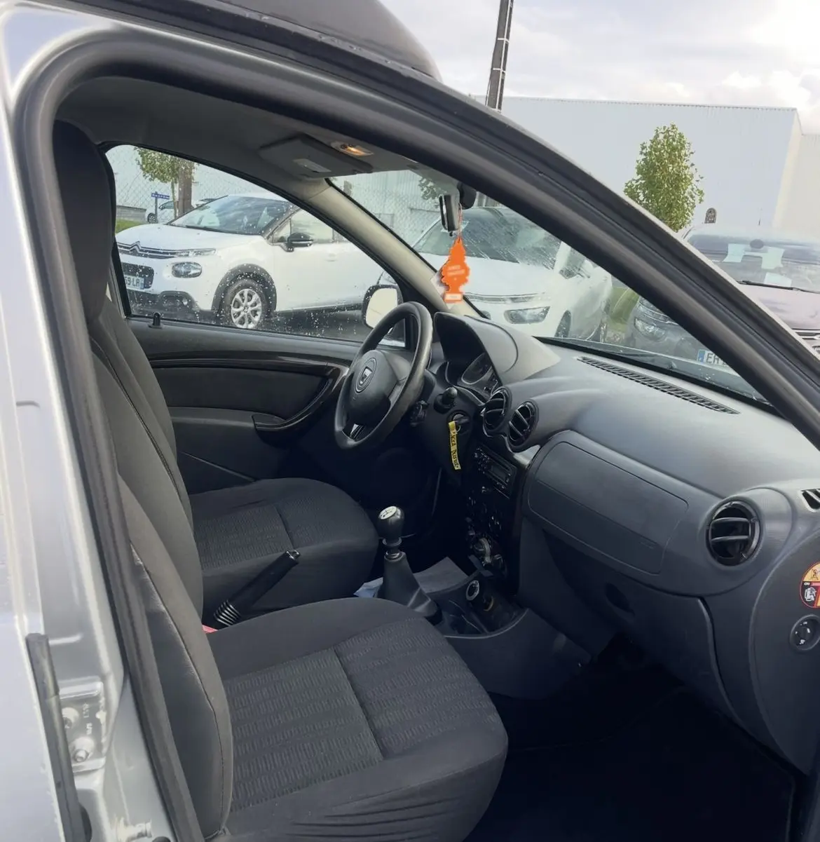 Vue intérieure côté conducteur du Dacia Duster gris 2011, montrant les sièges noirs et le tableau de bord simple avec boîte manuelle.