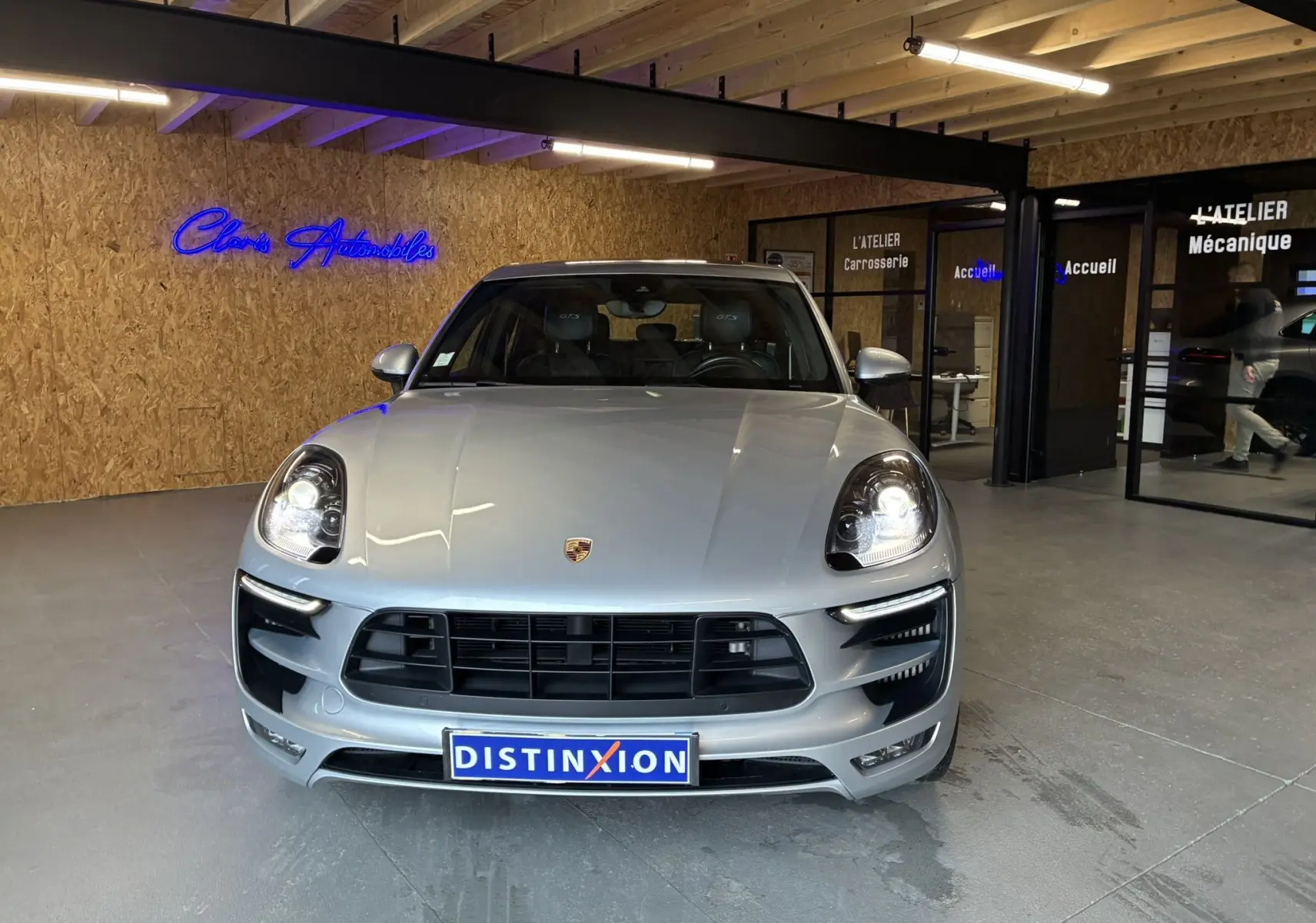 Vue frontale d'un Porsche Macan GTS argenté avec phares allumés, calandre noire et plaque Distinxion.