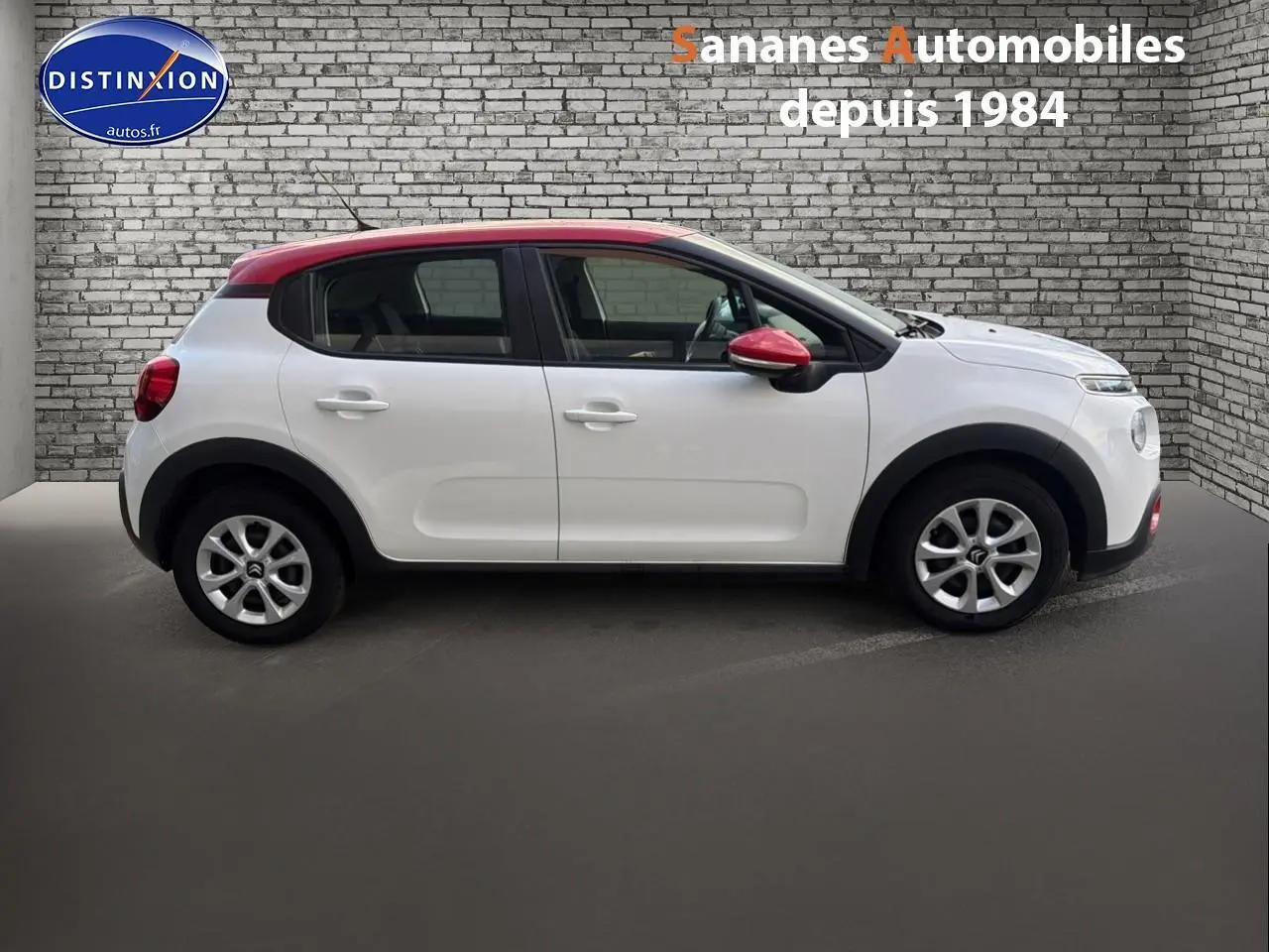 Profil droit d'une Citroën C3 blanche avec toit et rétroviseur rouge, sur fond de mur en briques grises.